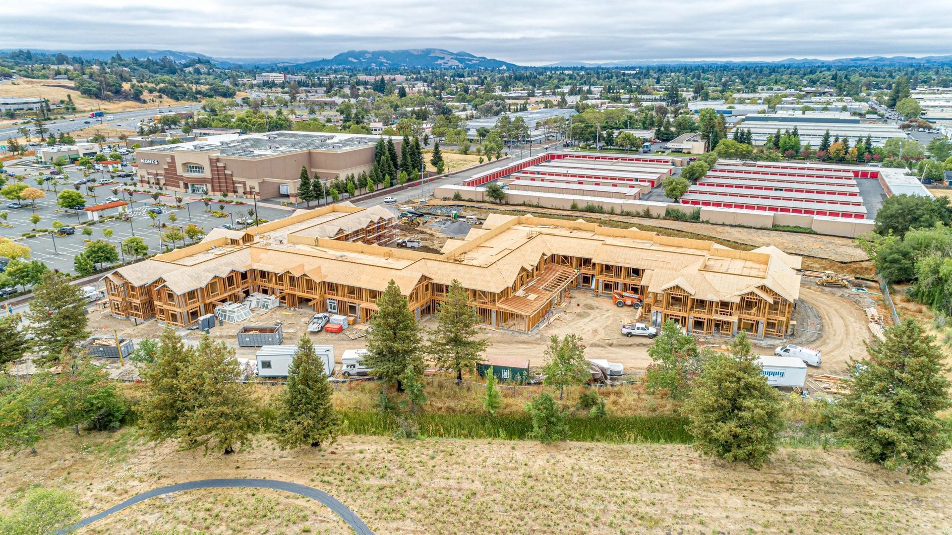 Aerial view of a wooden residential building frame under construction, surrounded by trees, parking lots, and a town.