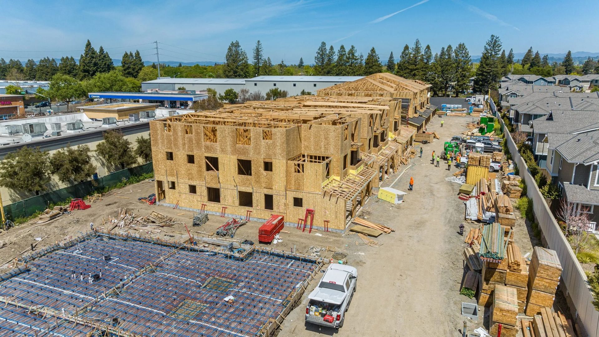 An aerial view of a construction site with a wooden apartment frame, foundation work, and a parked white truck.