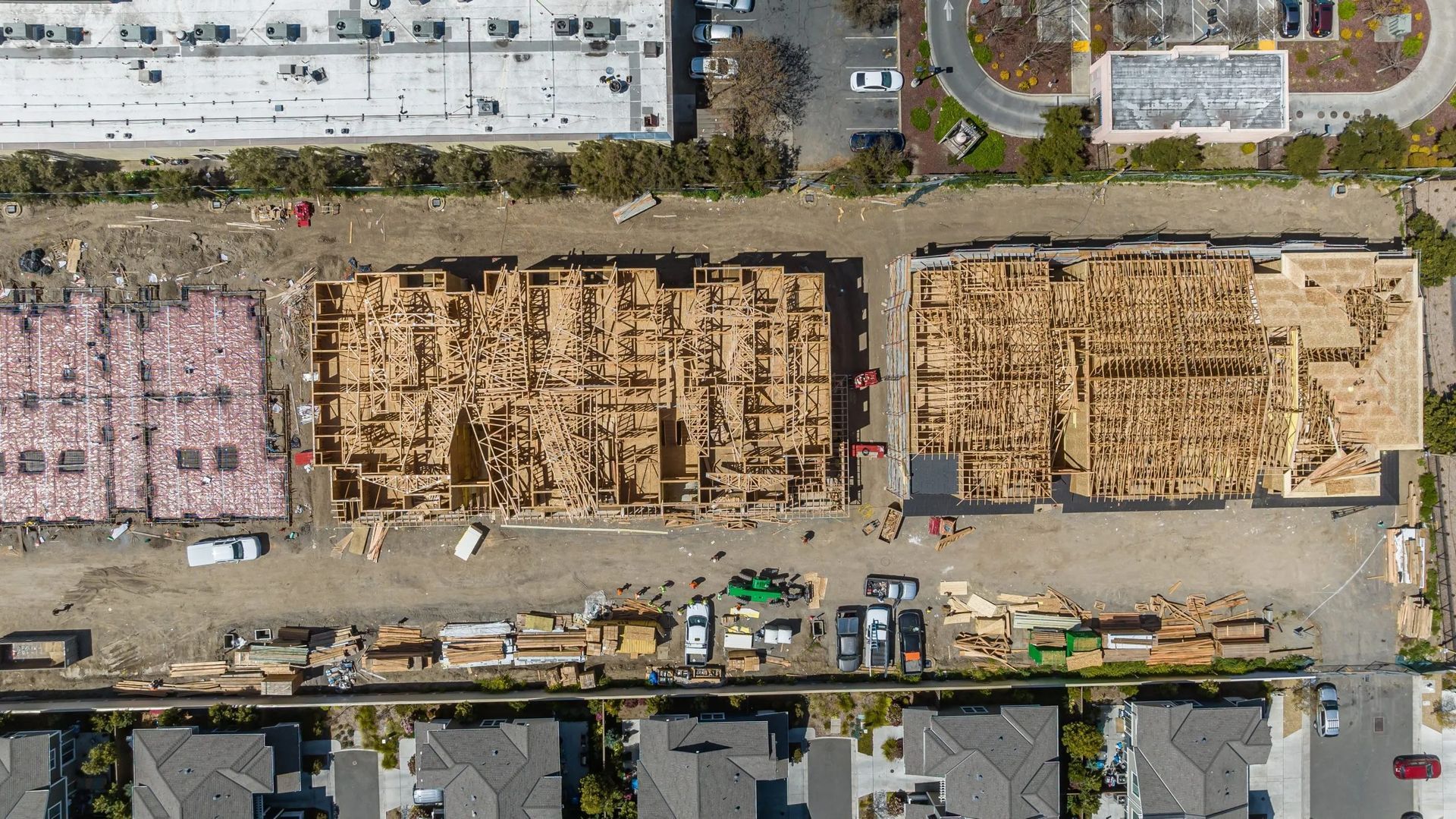 Aerial view of a construction site with lumber piles, equipment, and adjacent buildings and parking lots