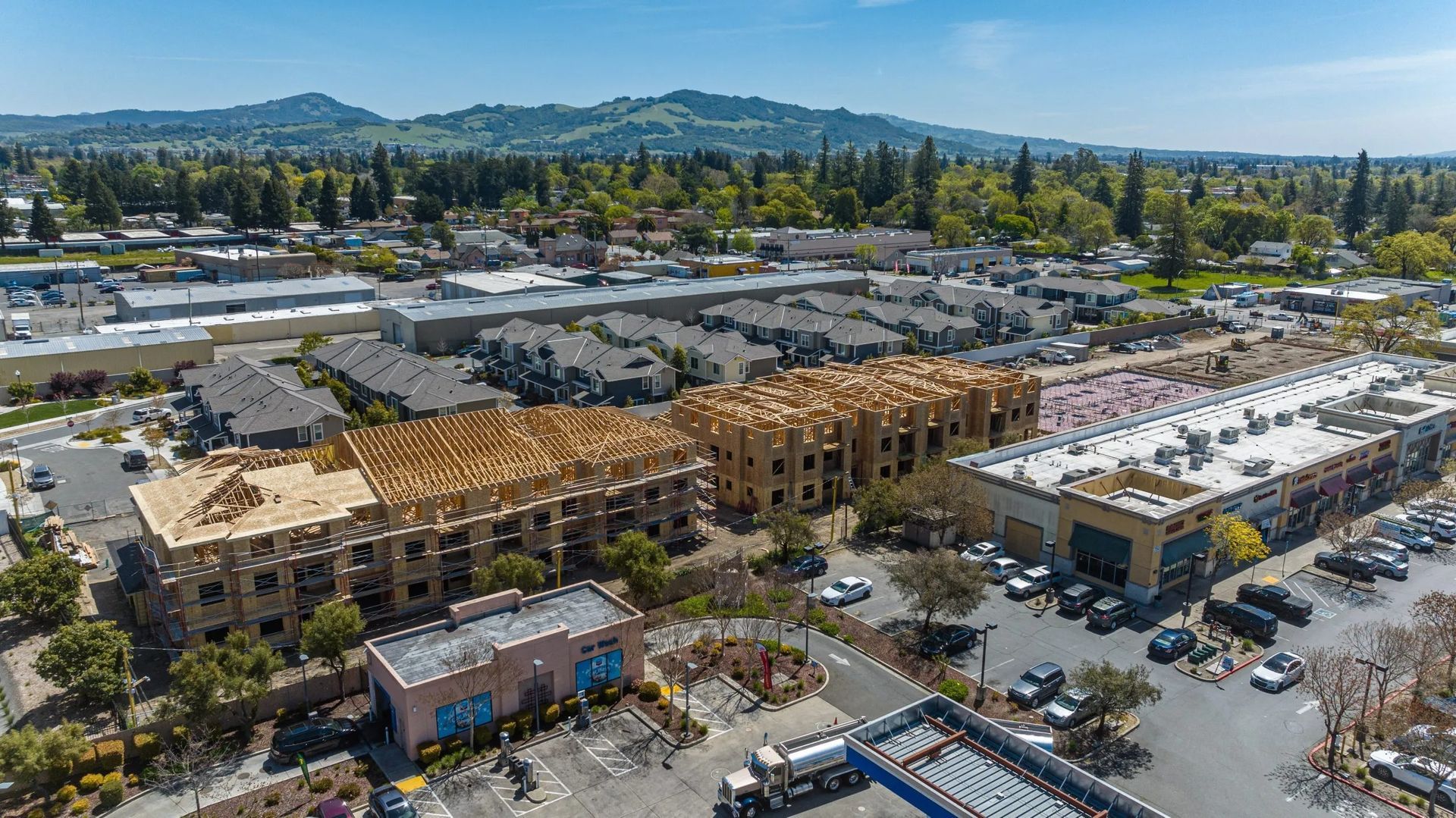 Aerial view of a residential construction site with wooden frameworks under a clear sky, surrounded by buildings and cars.