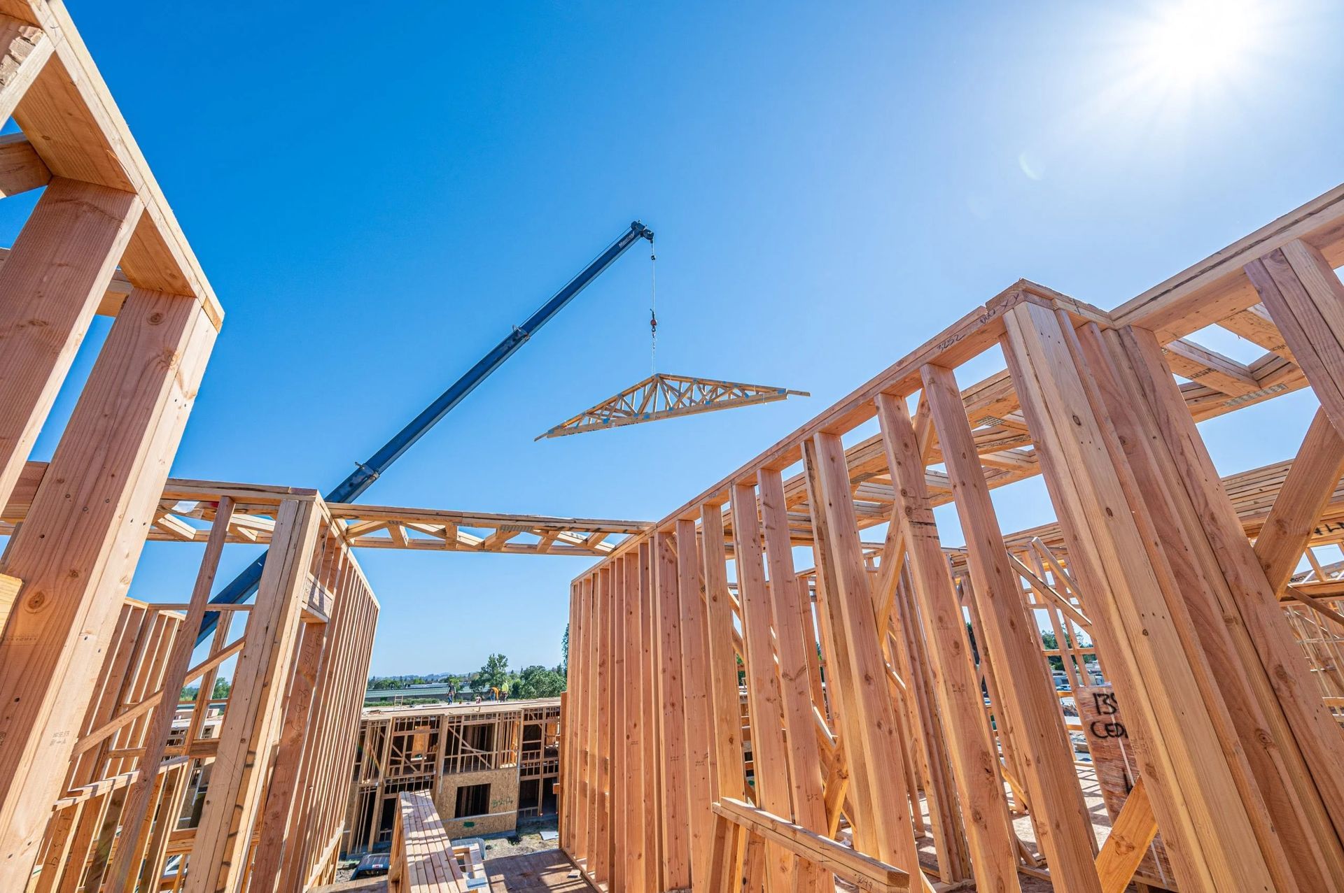 Wood-framed house under construction with crane lifting roof trusses against a bright blue sky