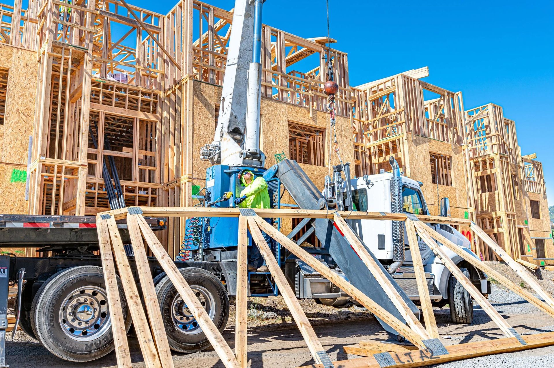 A crane truck parks in front of a wooden building frame under construction against a clear blue sky.