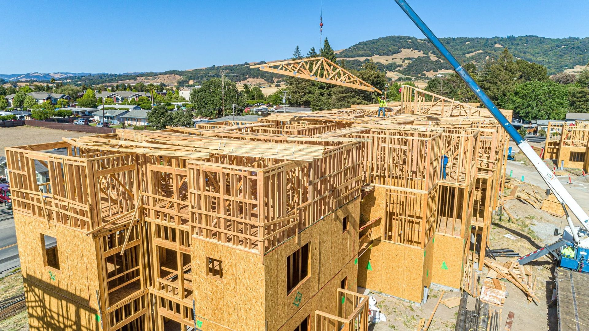 A crane hoists a wooden roof truss over a multi-story apartment building under construction on a sunny day.