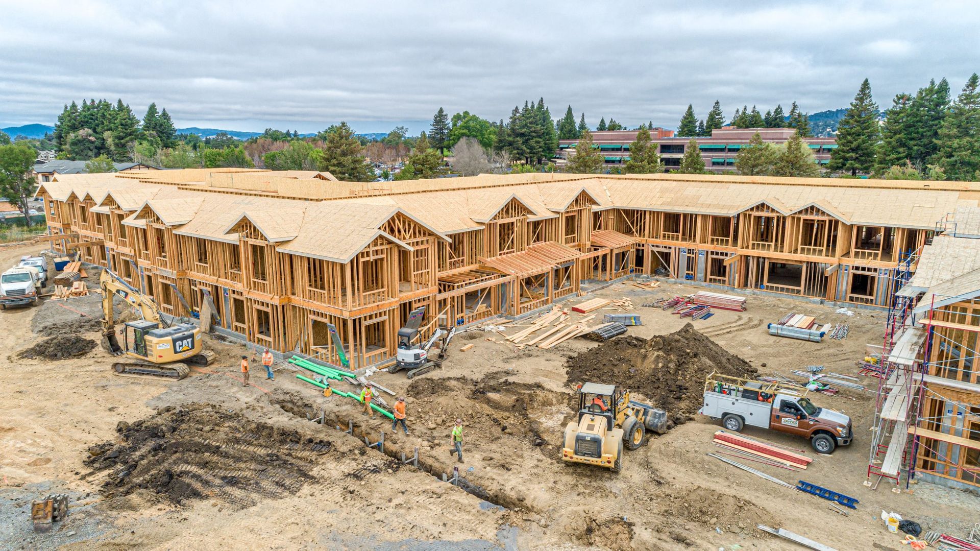 Construction site of a large, wooden-framed apartment building with excavators, a service truck, and dirt mounds.