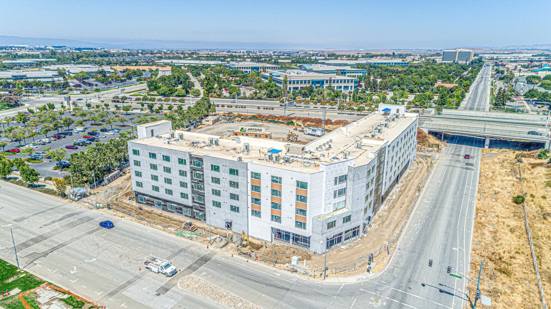 An aerial view of a multi-story, white apartment building under construction, surrounded by roads and sparse greenery.