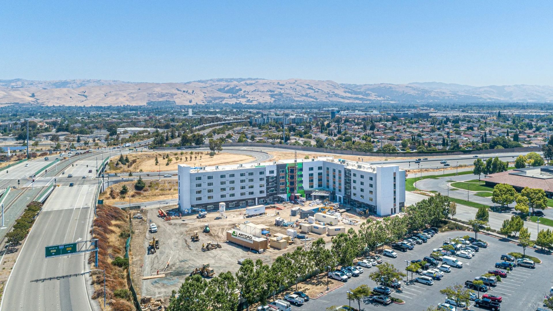 An aerial view of a multi-story hotel under construction, next to a highway and a large parking lot, under a clear sky.