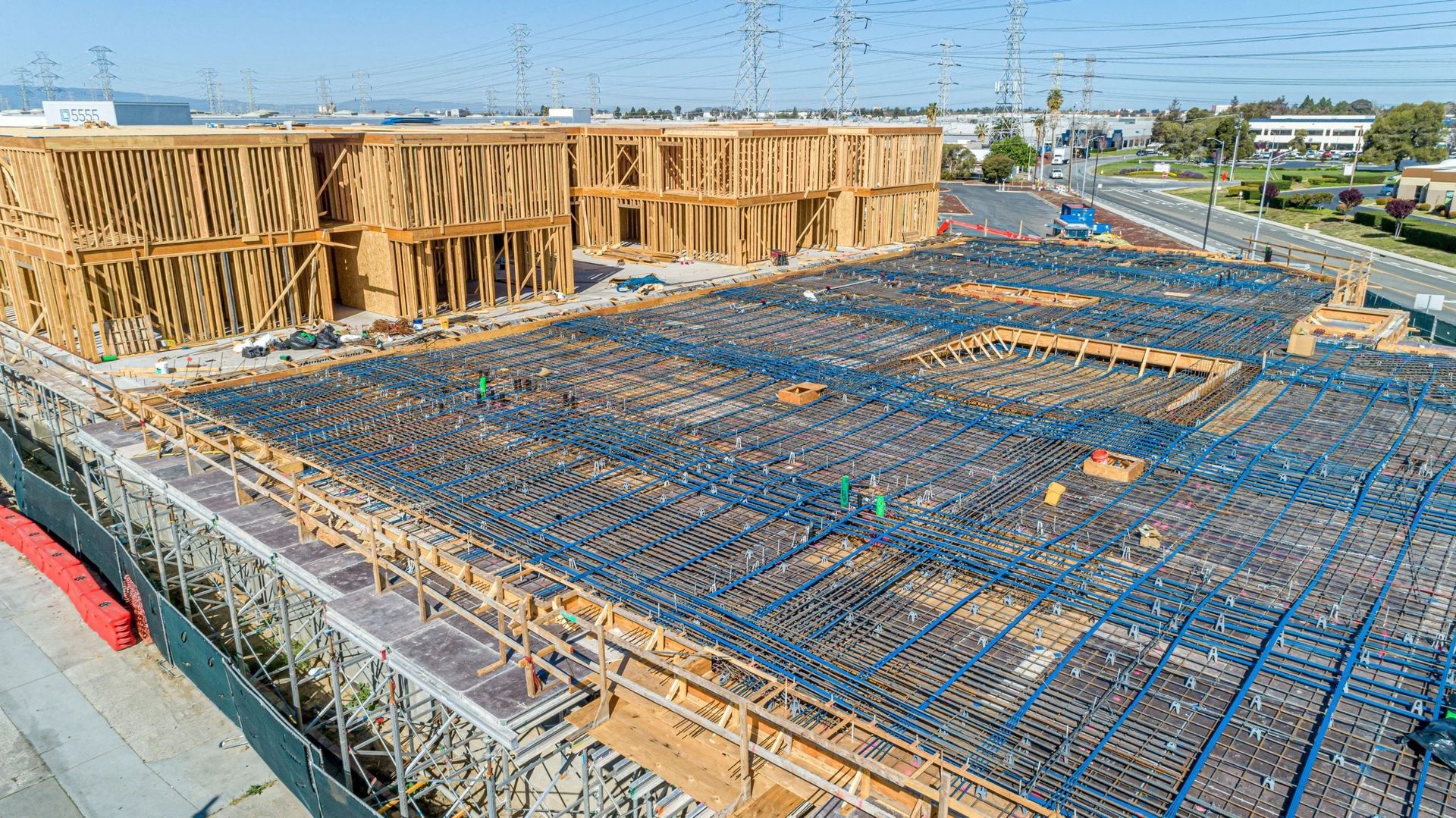 An elevated view of a construction site showing a large rebar grid on the ground and partially built wooden structures.