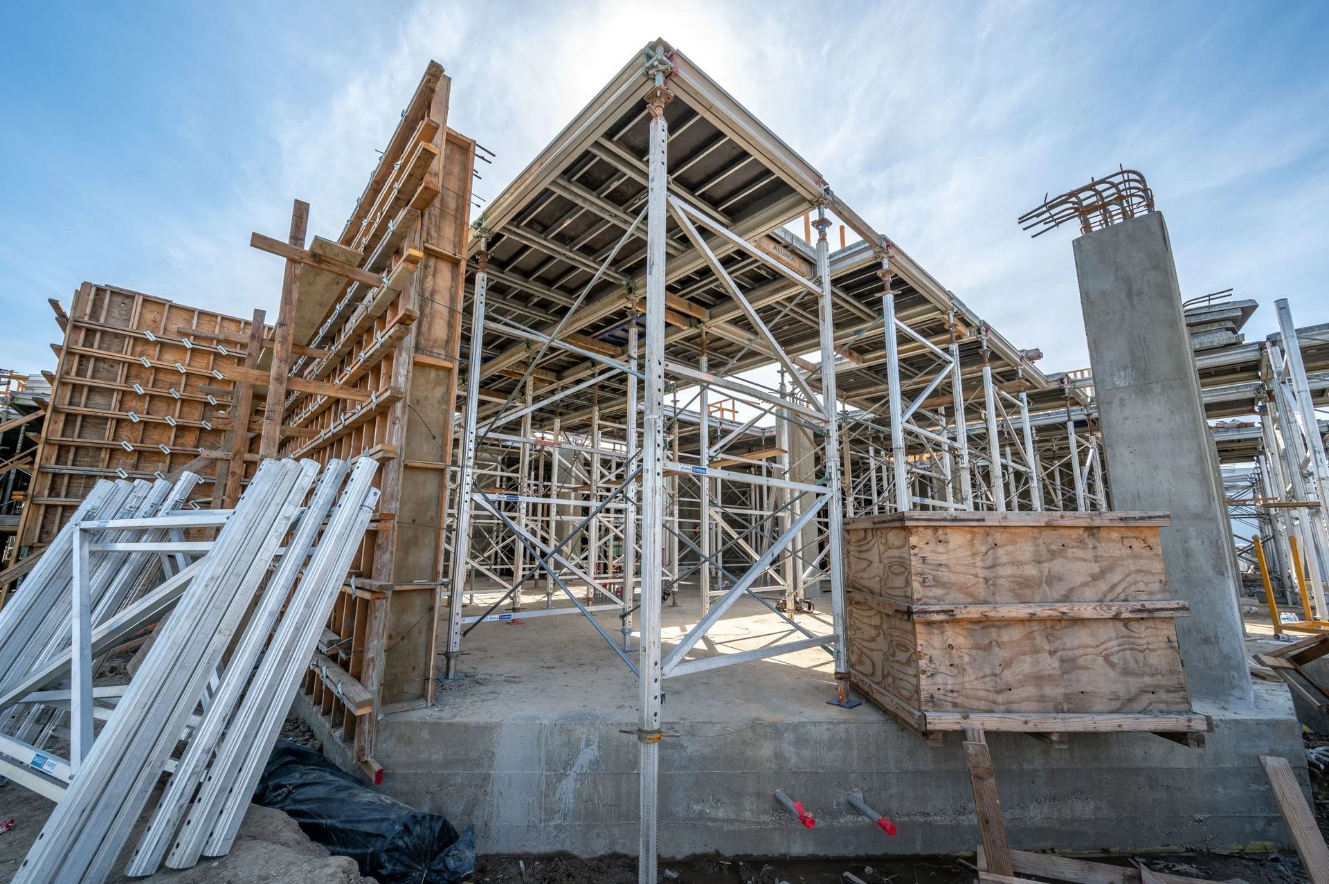 A construction site featuring metal scaffolding and wooden forms supporting a concrete slab under a bright blue sky.