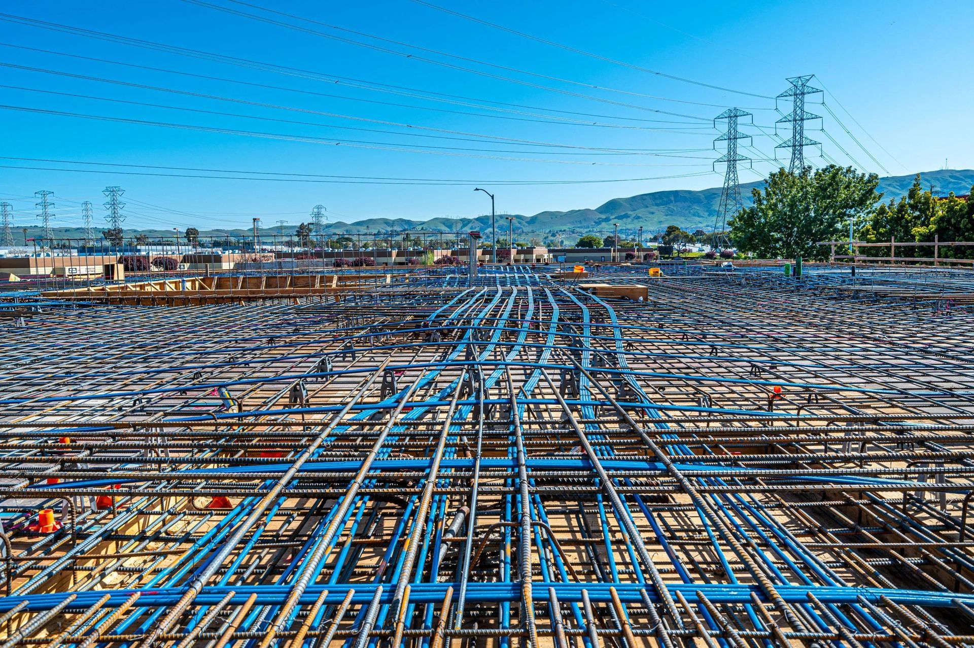 A construction site shows a dense grid of blue electrical conduits and steel rebar spread across a large concrete foundation.