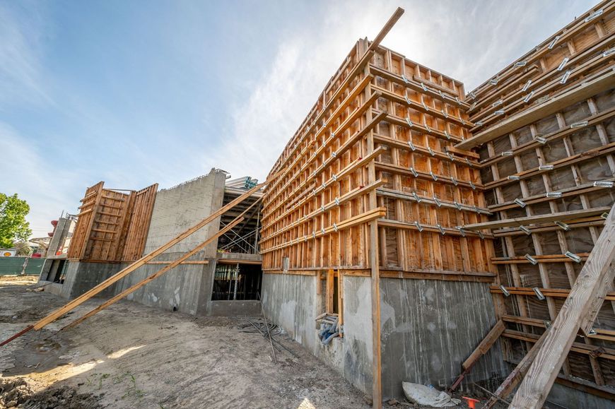 A construction site showing concrete walls under assembly with large wooden bracing and formwork against a blue sky.