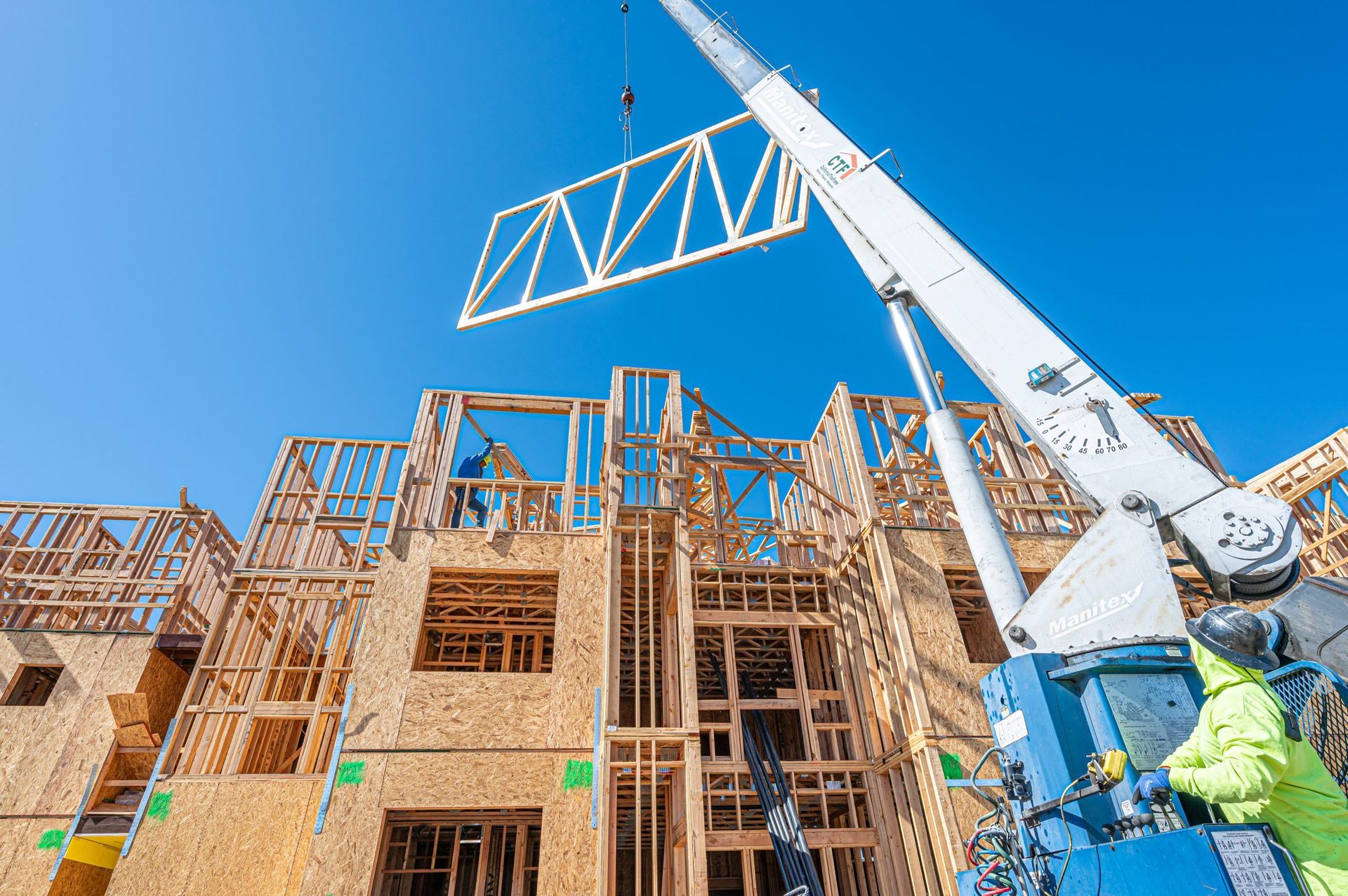 A crane lifting a wooden wall frame to the upper level of a building under construction against a clear blue sky.