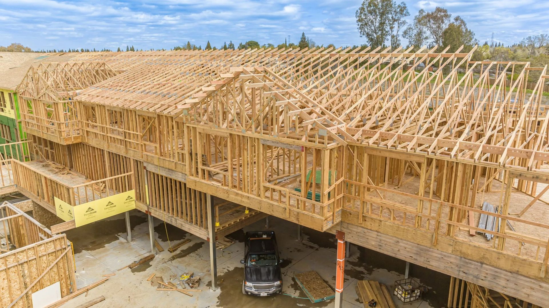 An aerial view of a multi-story wooden house frame under construction, with a black truck parked underneath the structure.