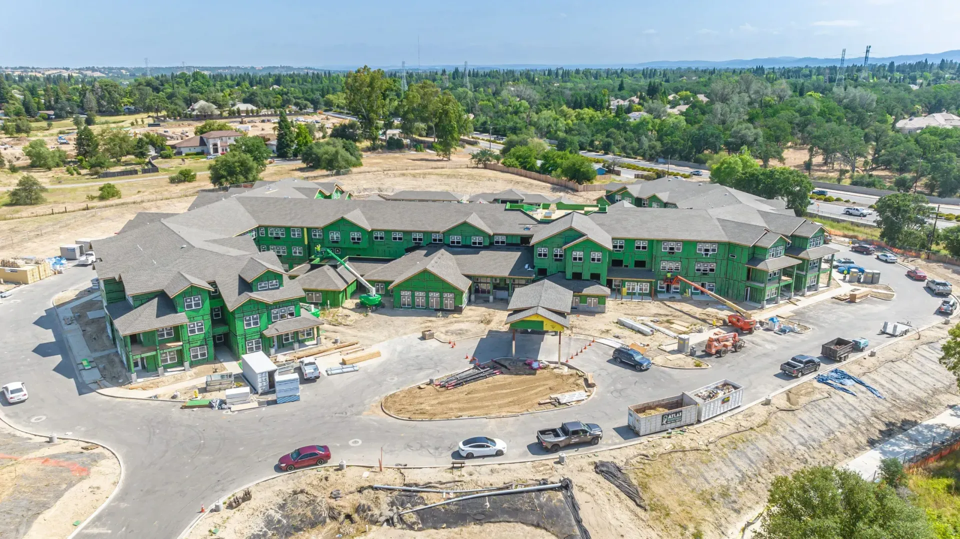 Aerial view of a large green-roofed building complex on a dusty construction site with vehicles and trees nearby