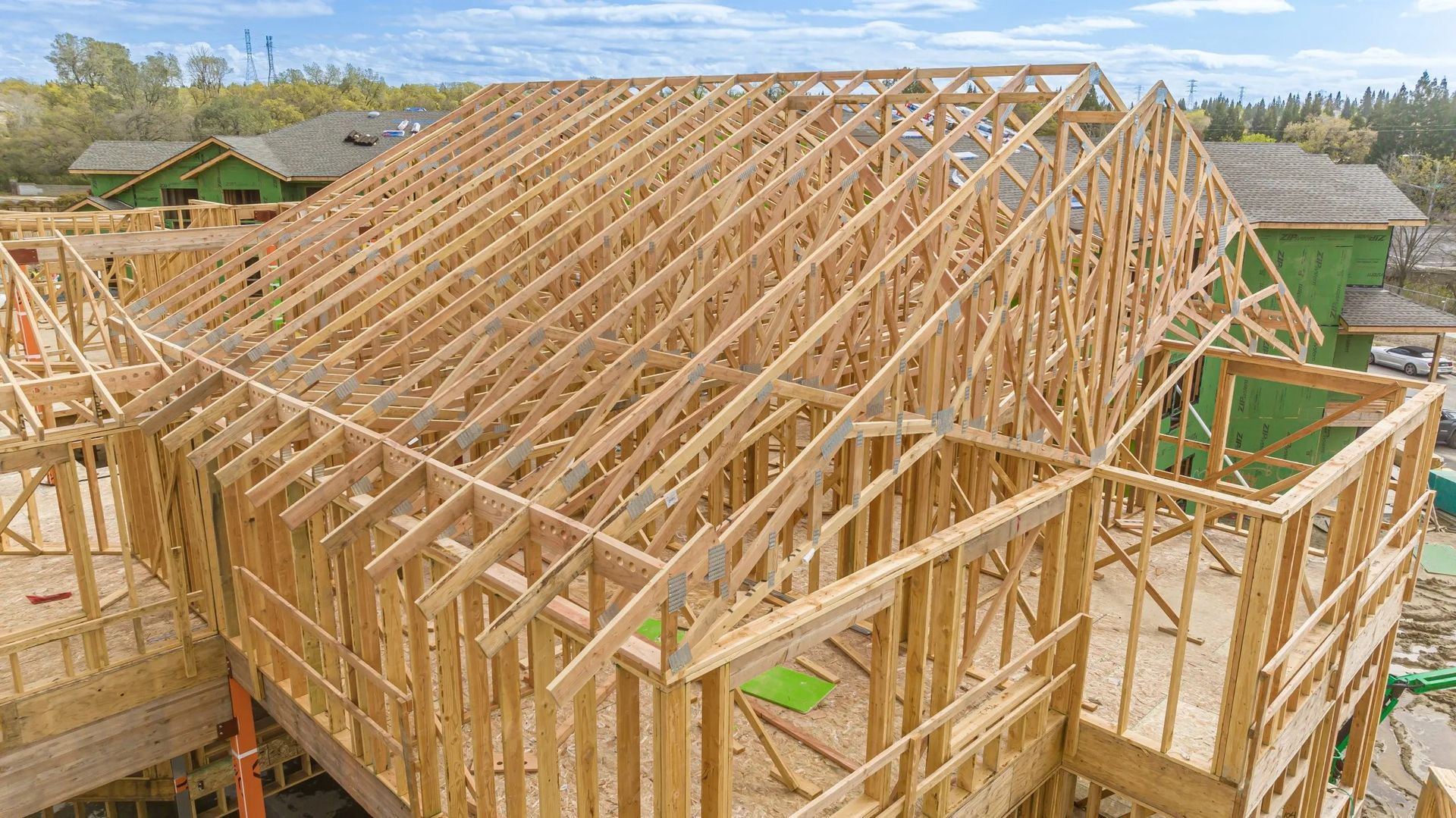 A high-angle view of a wooden house frame under construction against a blue sky with surrounding neighborhood homes.