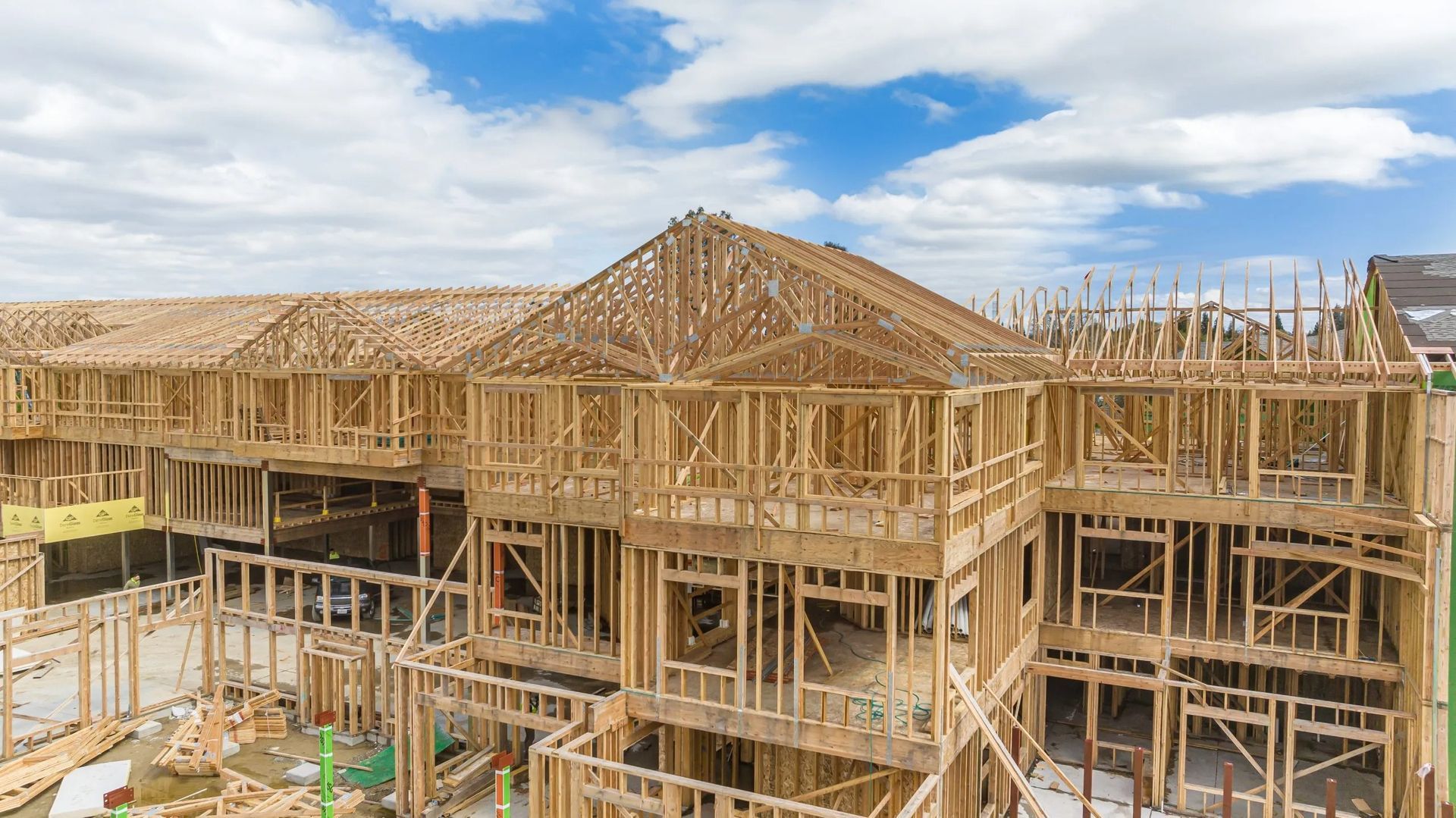 A low-angle view of several multi-story wooden house frames under construction against a partly cloudy blue sky.