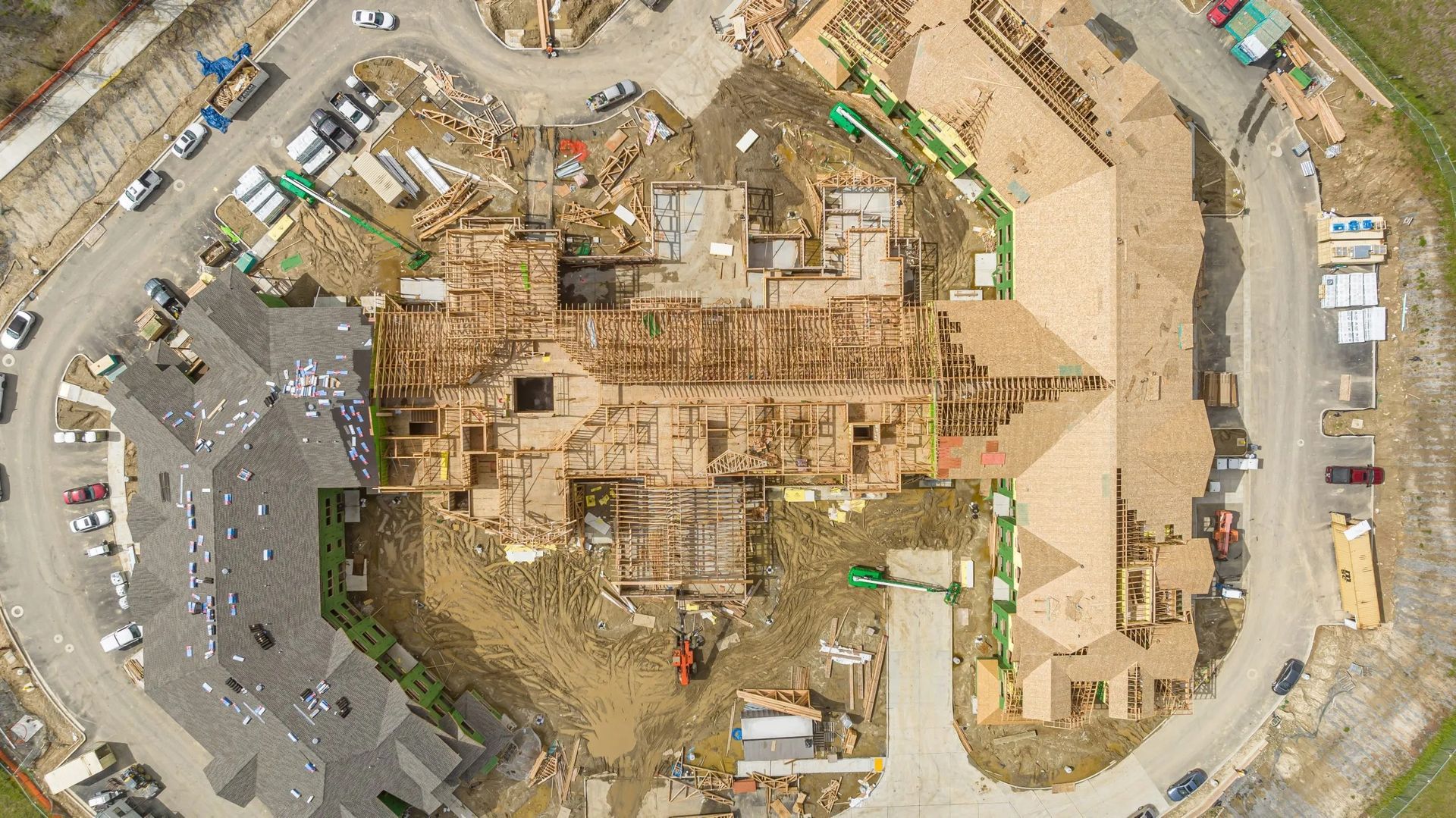 Aerial view of a residential construction site with multiple buildings, wooden roof framing, and surrounding roads.