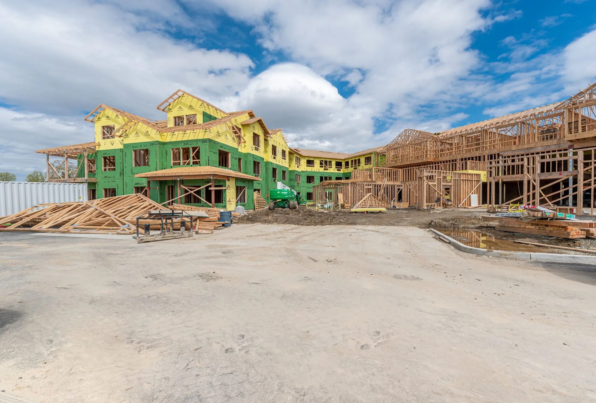 Partially built houses under construction, with green siding and wooden framing on a dirt lot under a cloudy sky