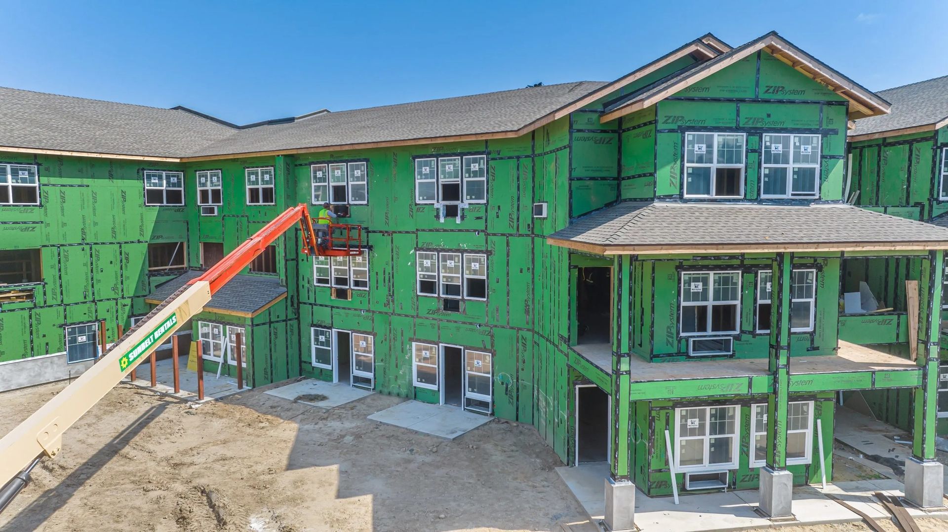 An unfinished apartment complex under construction with green exterior sheathing, an orange lift, and a blue sky.