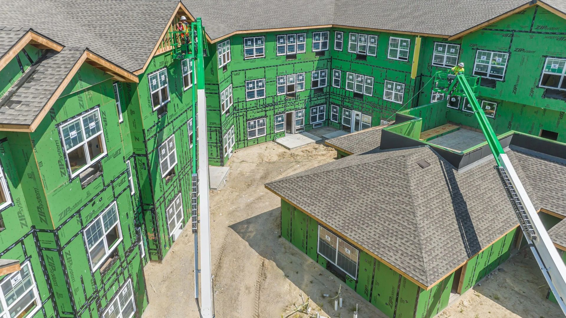 An aerial view of a multi-story apartment building under construction, featuring green sheathing and roof shingles.