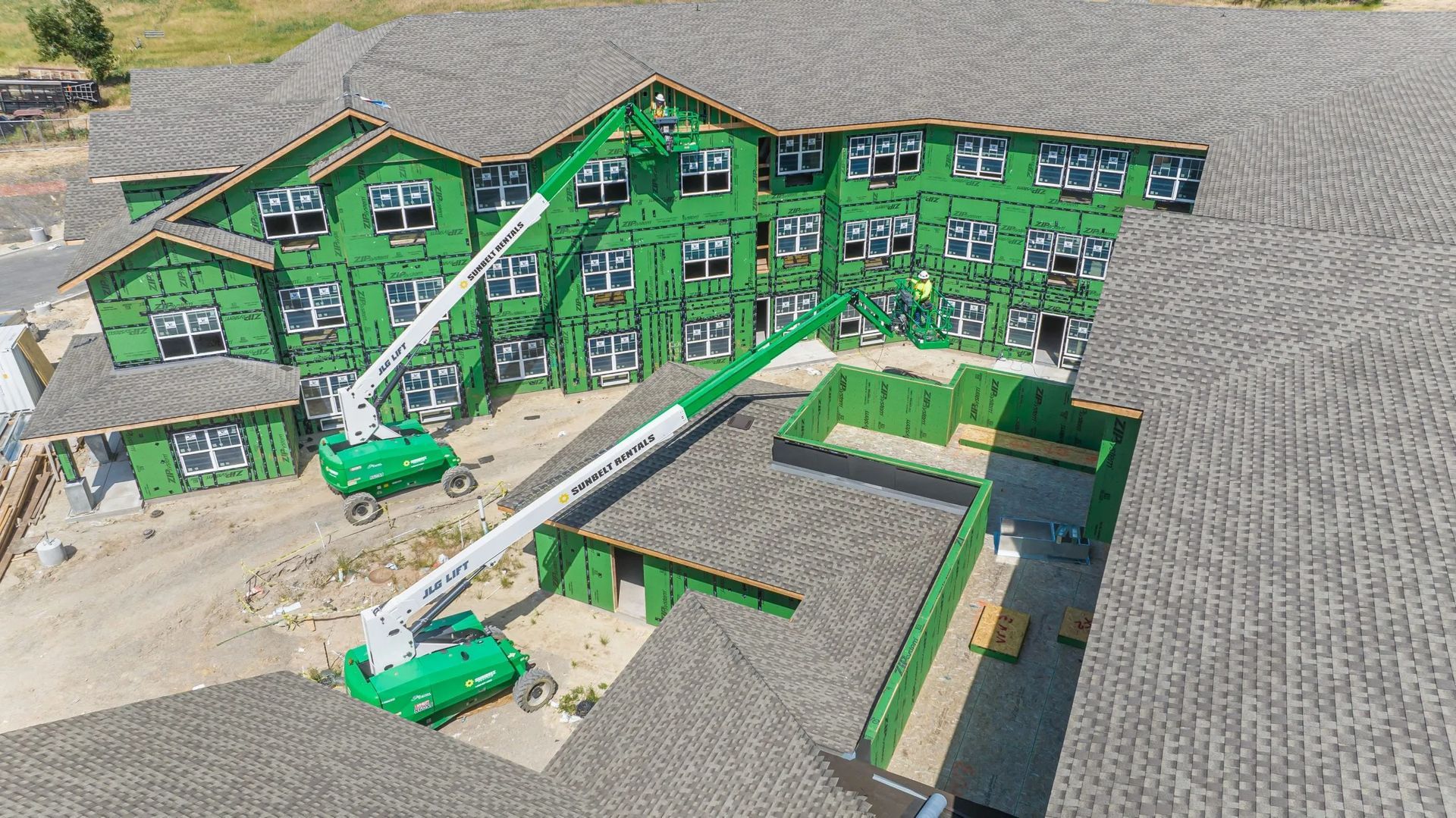 An aerial view of a multi-story building under construction with green sheathing and two green boom lifts on the site.
