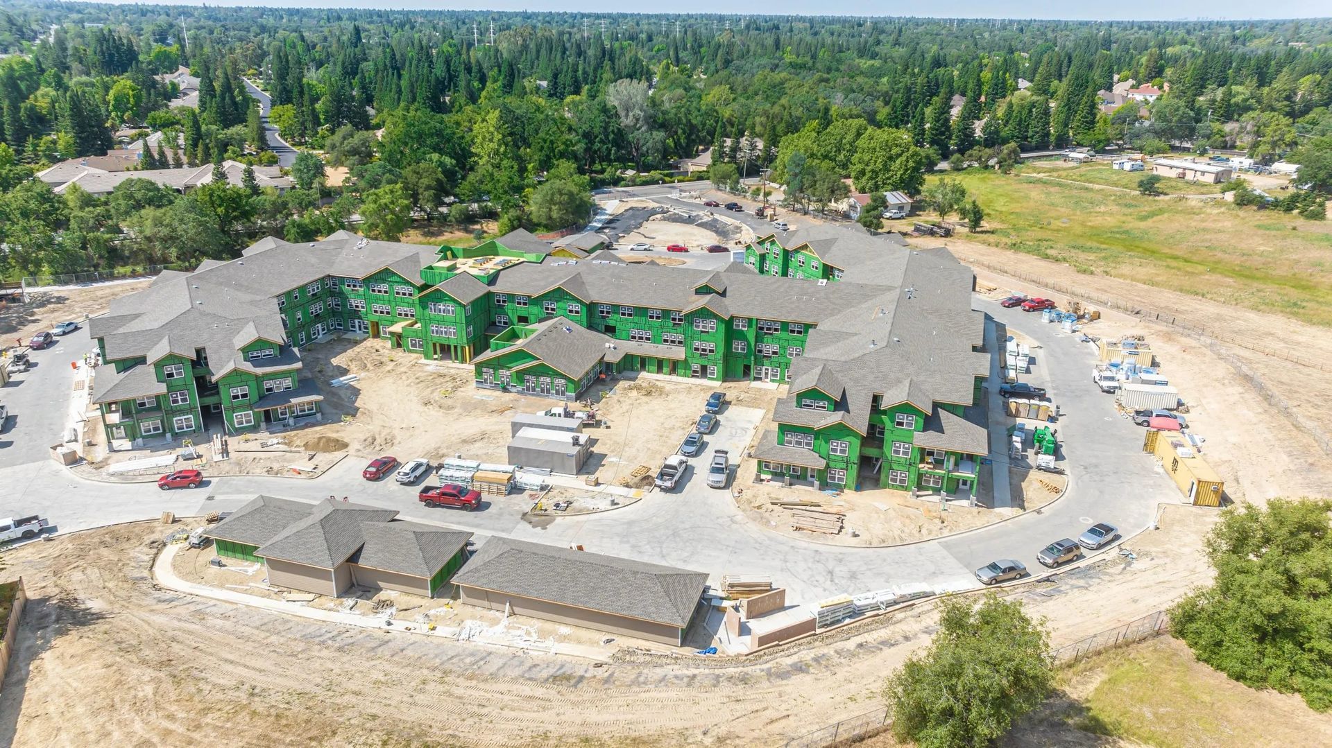 Aerial view of a multi-story apartment complex under construction with green sheathing, parking lots, and dirt surroundings.