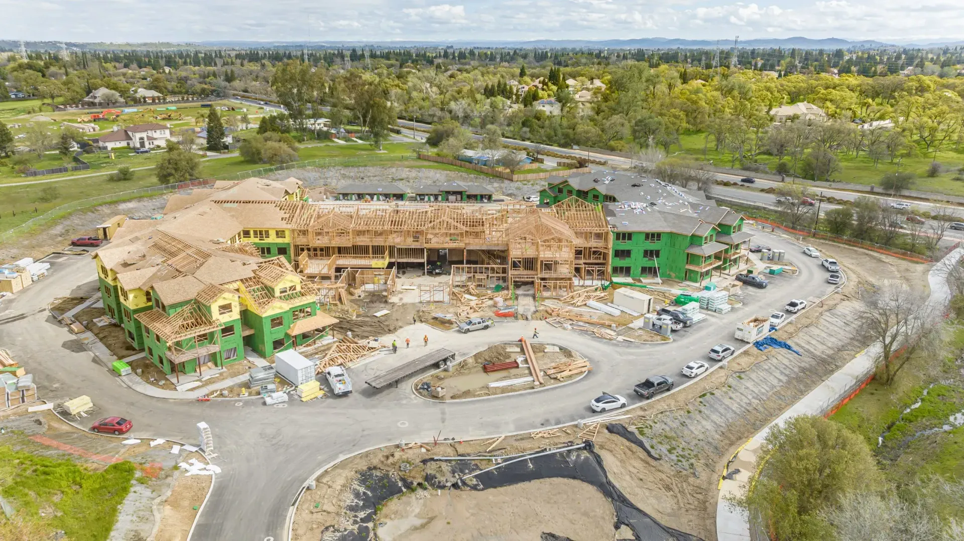 Aerial view of a large construction site with a multi-story building, machinery, and dirt roads surrounded by trees.