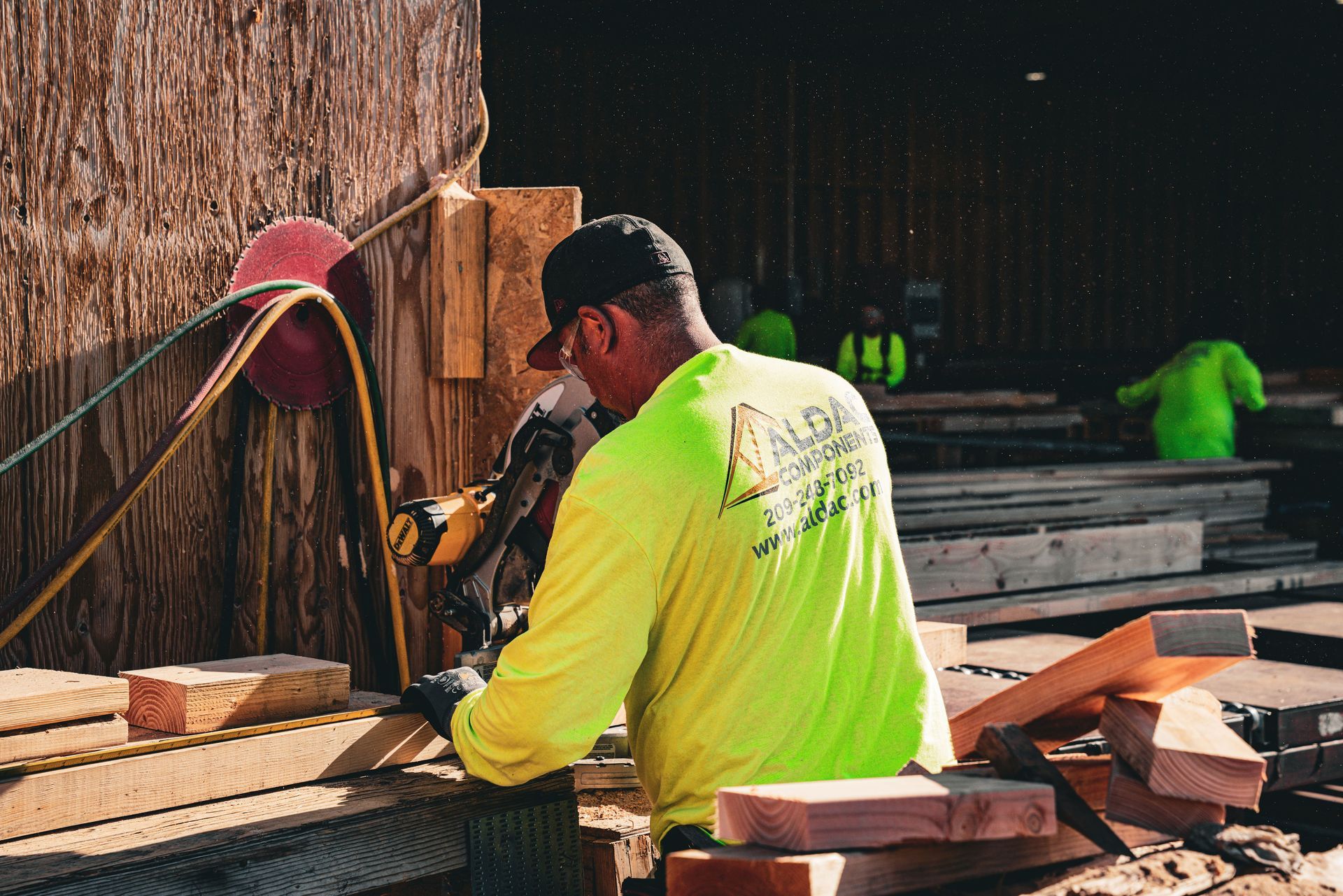 Worker in neon yellow jacket cutting wood at an outdoor workbench with lumber stacked nearby