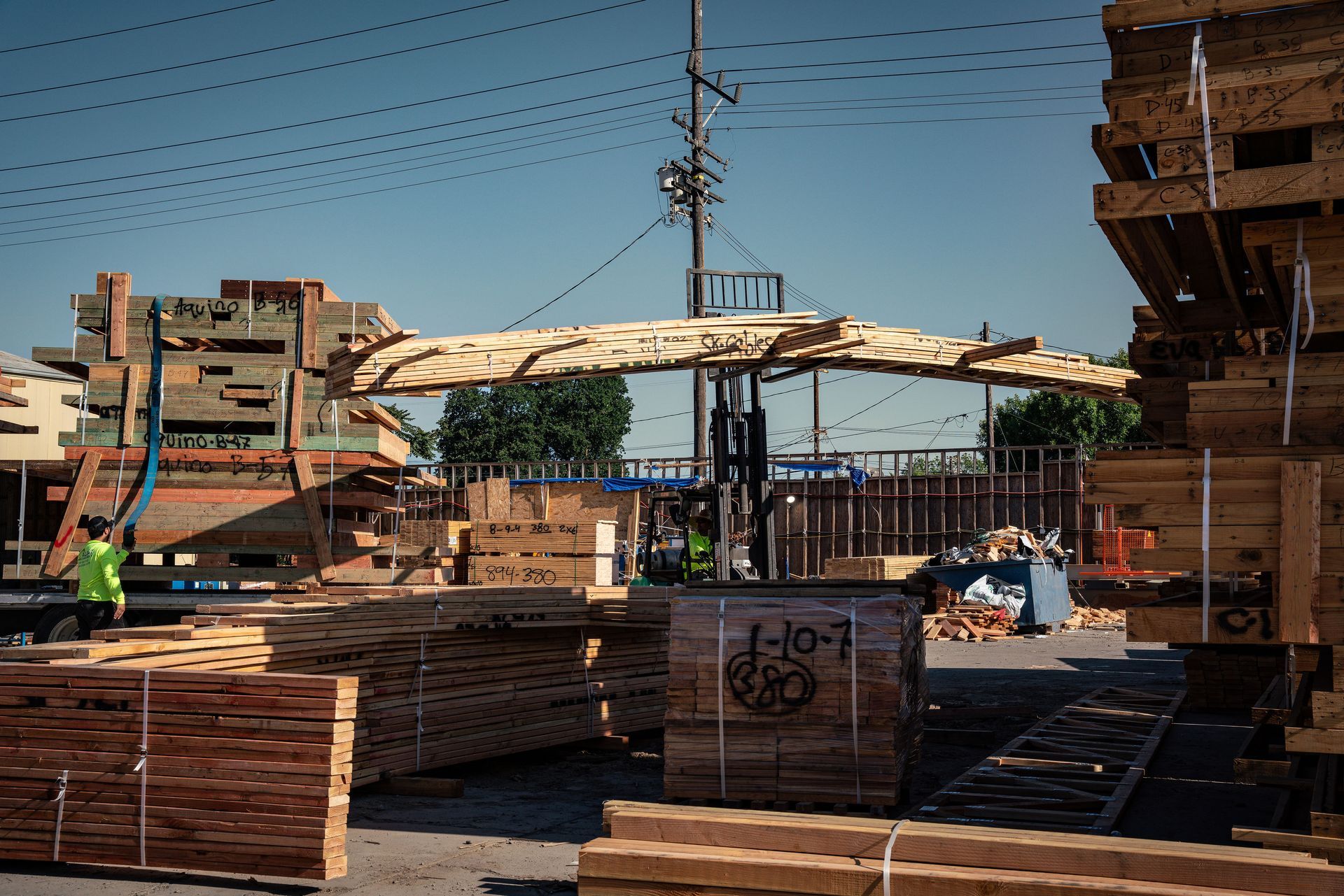 Construction site with stacked lumber and workers under a canopy beside a tall building.