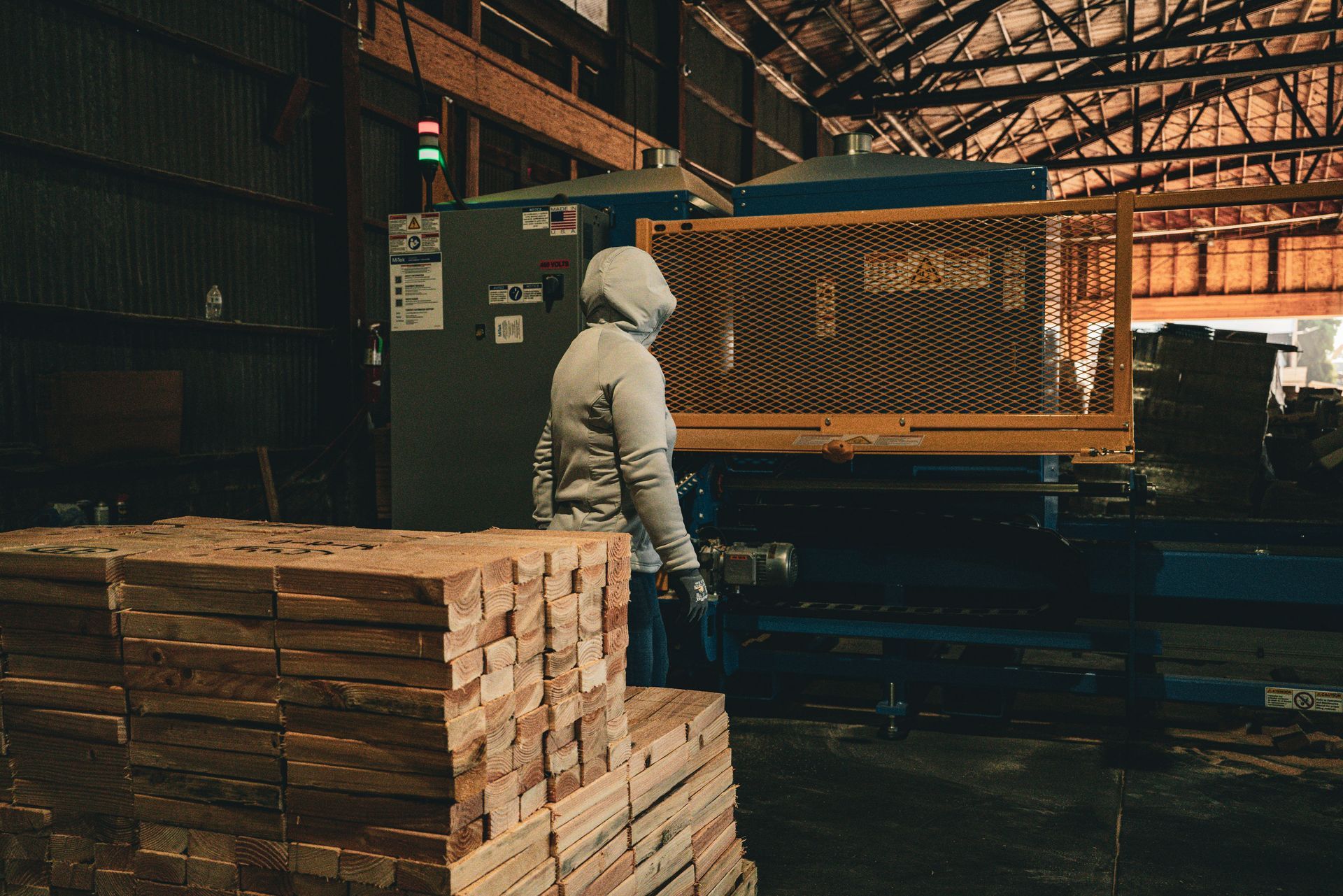 Worker in a white hoodie stands by stacked wood in a dim warehouse near a large machine.
