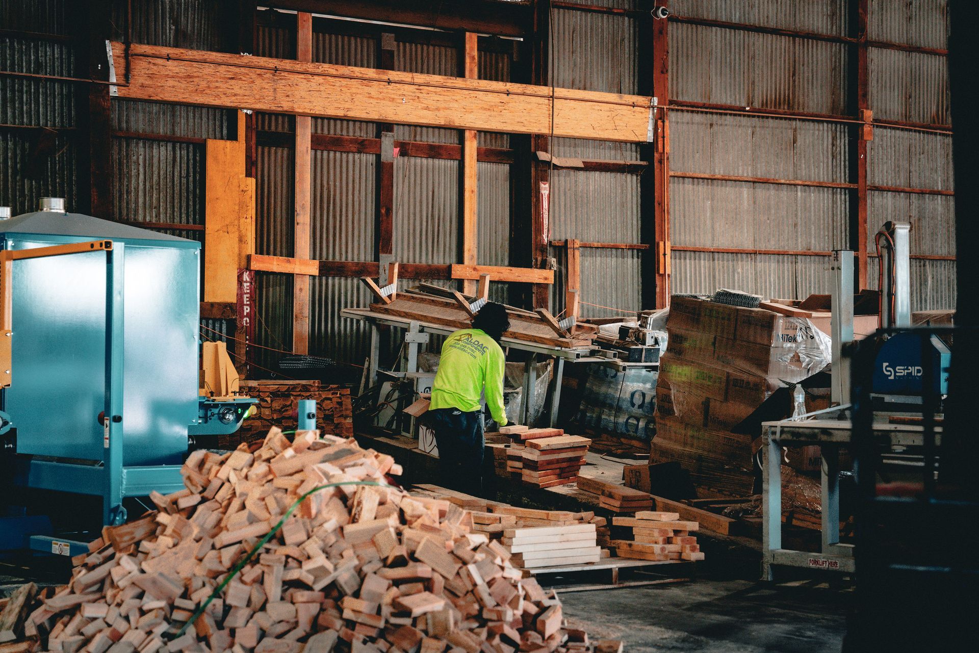 Worker in a neon vest sorting lumber inside a woodshop with piles of cut wood and sawdust