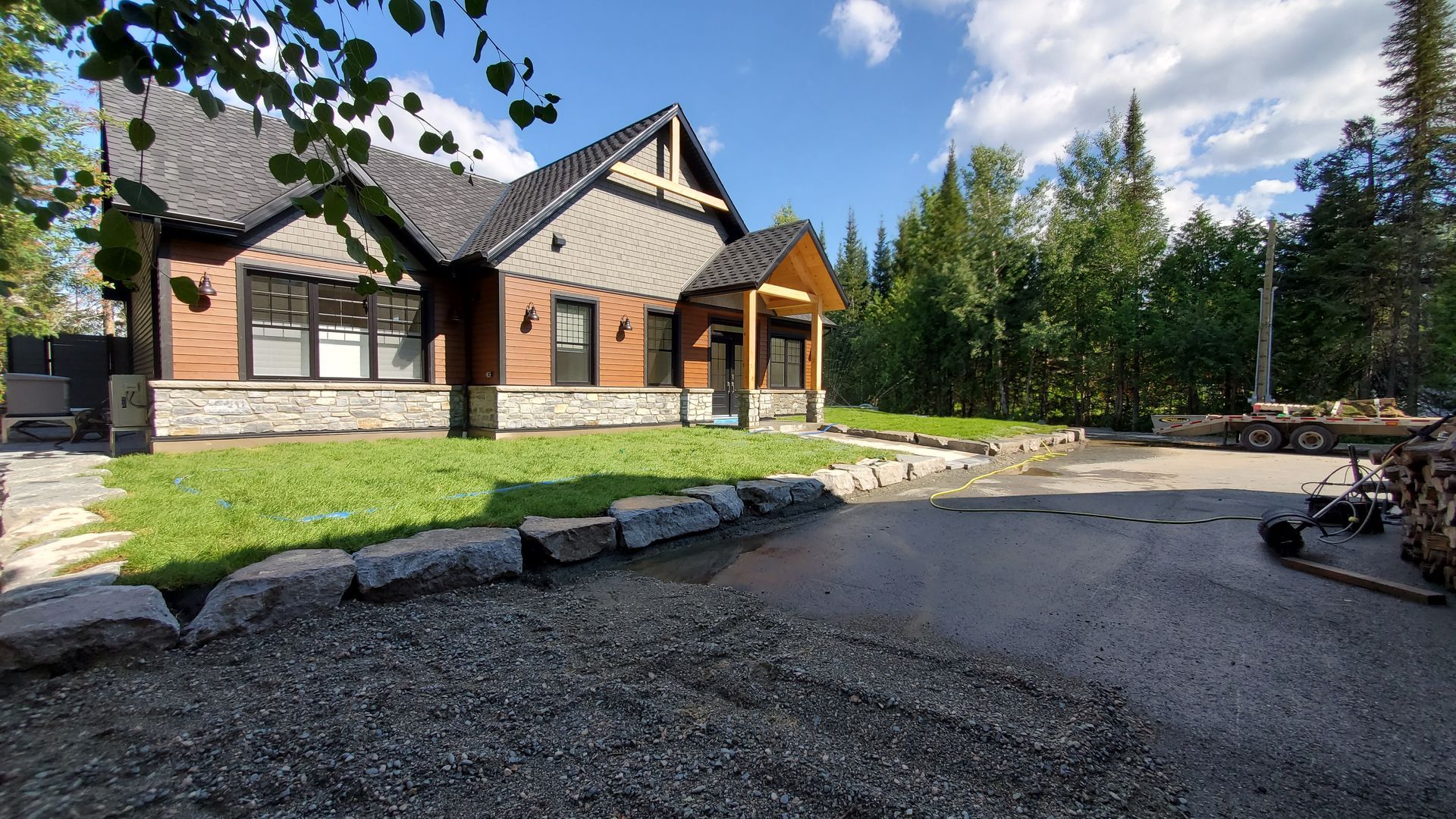 Maison de campagne avec façade en bois et pierre, située sur une pelouse verdoyante avec une allée en gravier. Arbres verdoyants en arrière-plan sous un ciel bleu.