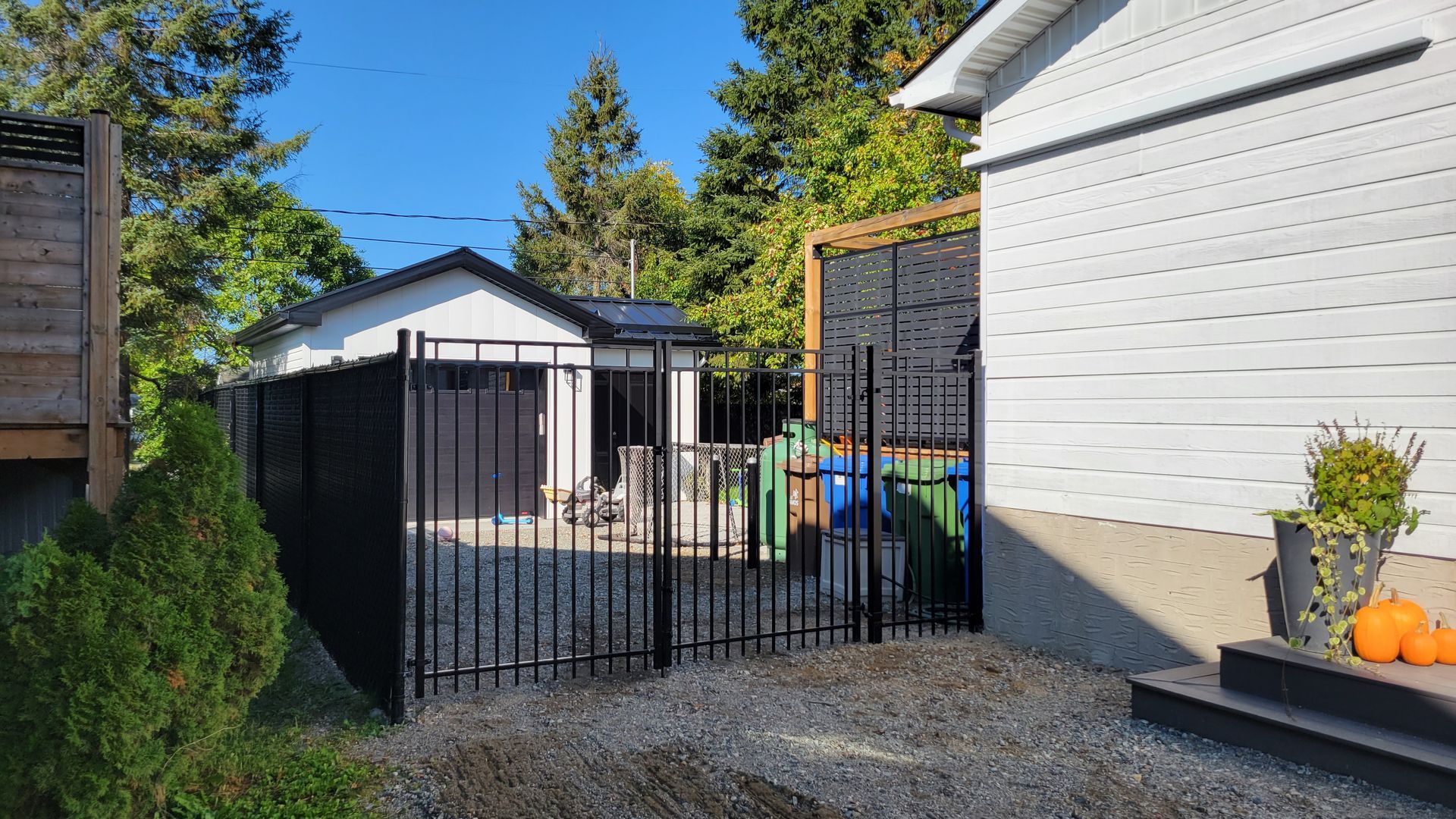 Clôture métallique noire devant un garage et poubelles vertes et bleues. Maison blanche à droite.