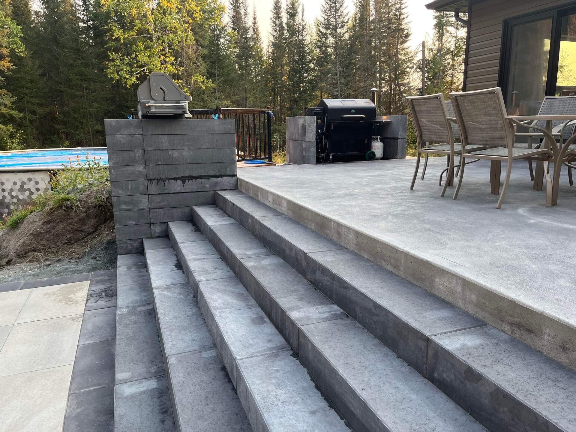 Jardin avec piscine rectangulaire, entourée d'une clôture noire, abri de jardin gris et maison grise sous un ciel dégagé.