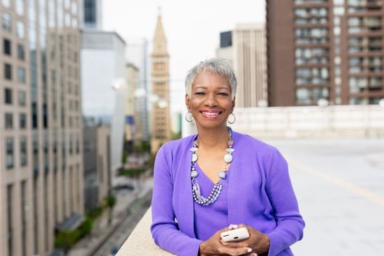 A woman in a purple sweater is standing on a balcony in front of a city.