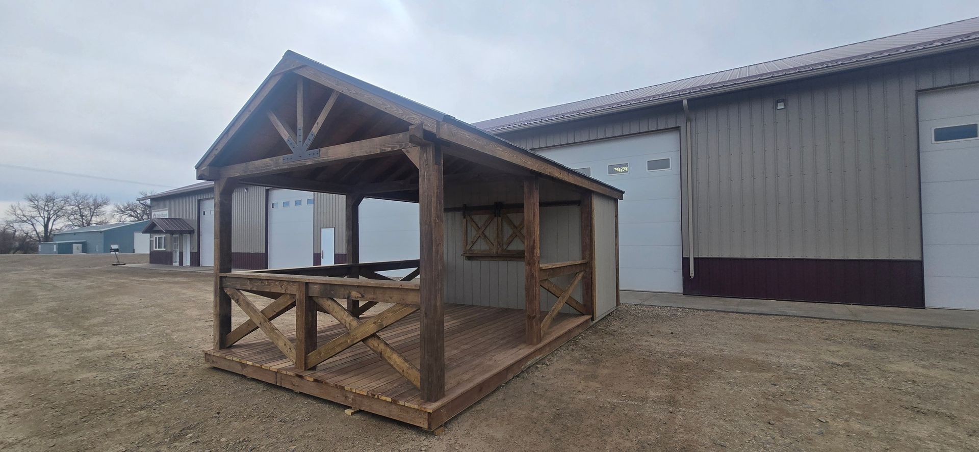A wooden, rustic outdoor structure with a covered porch and railing, situated on a dirt lot in front of a metal building.