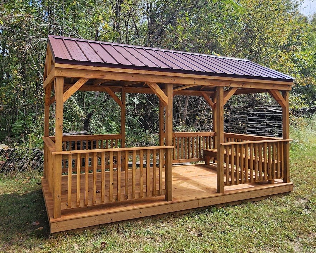 A wooden gazebo with a dark red metal roof and railings, situated in a grassy area with trees in the background.