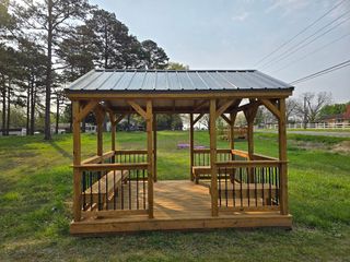 A wooden gazebo with a metal roof and built-in benches, set in a grassy outdoor area with trees in the background.