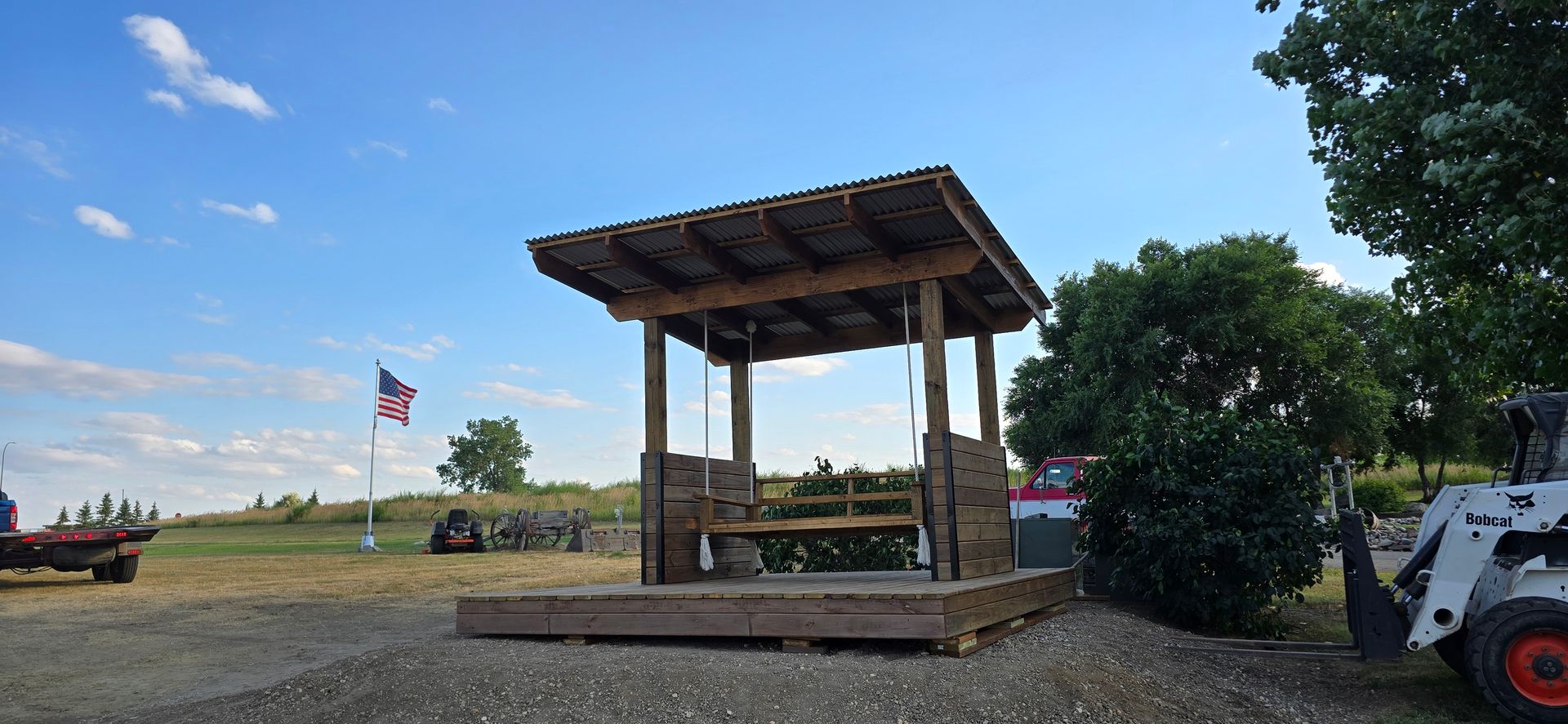 A wooden pergola with a built-in swing sits on a deck in a field under a bright blue sky, with an American flag nearby.