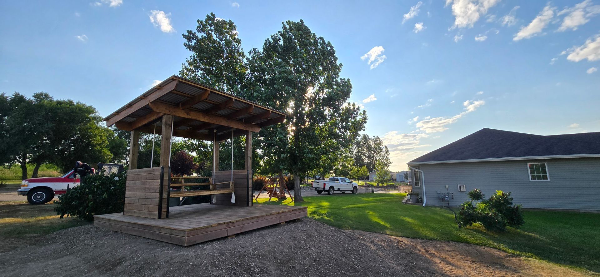 A wooden pergola with a built-in swing stands on a gravel patio in a grassy yard, with a house visible in the background.