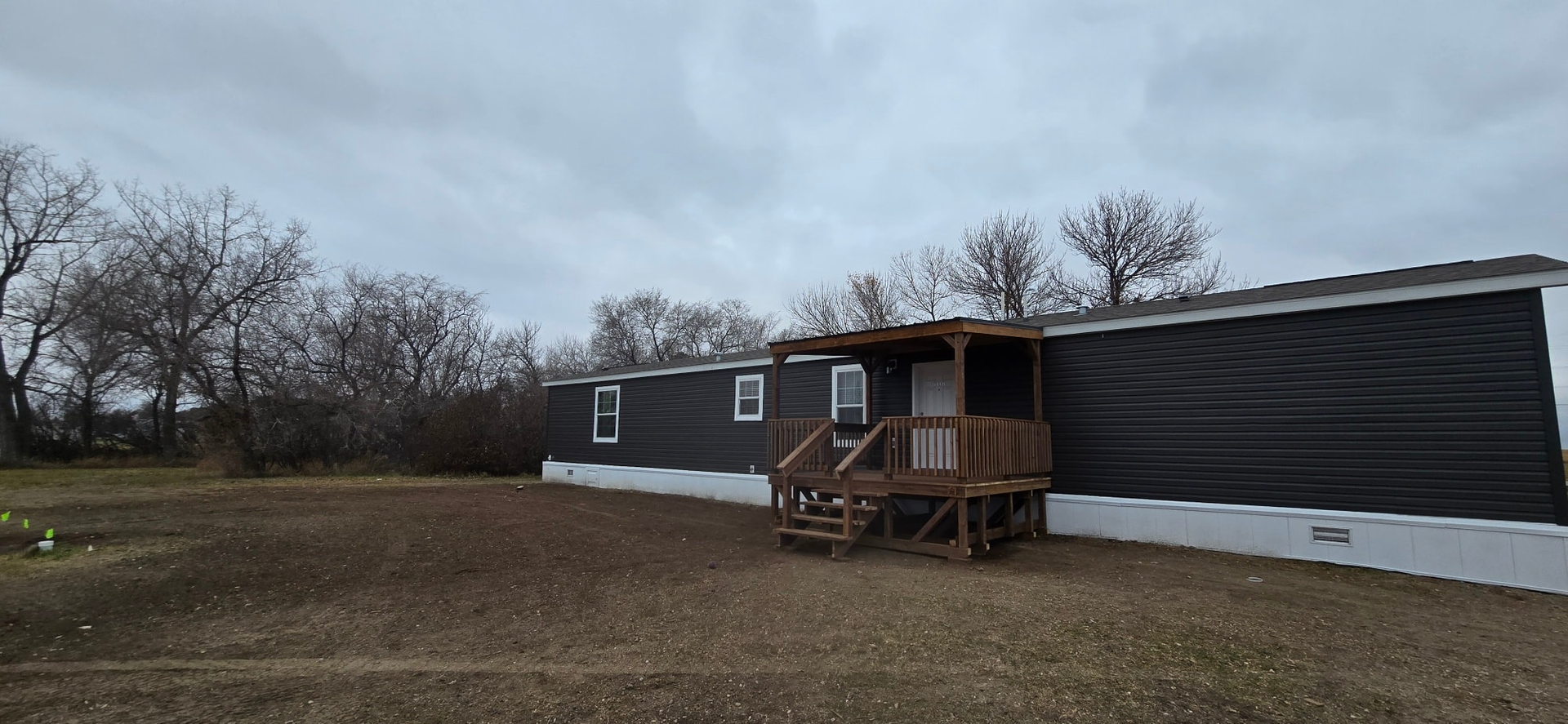 A dark-sided mobile home with a small wooden porch sits on a brown, grassy lot under an overcast sky.