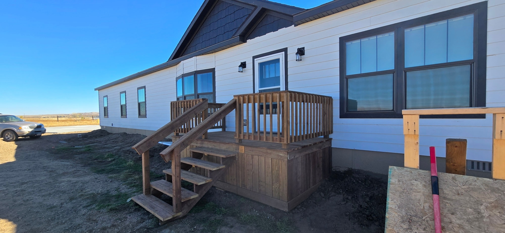 A partially finished house with white siding and dark trim, featuring a wooden deck and stairs under a clear blue sky.