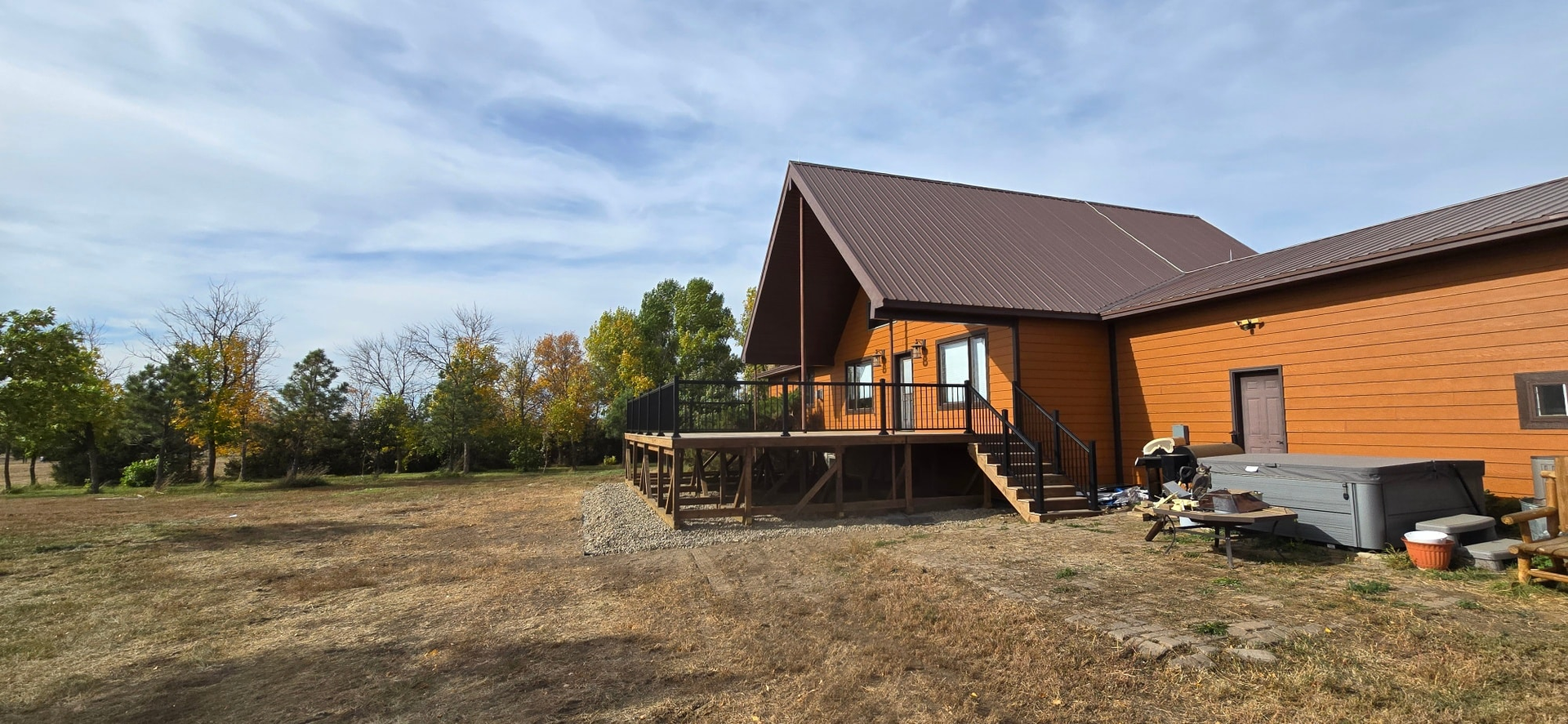 A rustic log cabin with a wooden deck and a hot tub on the patio, set in a dry, grassy field under a cloudy blue sky.