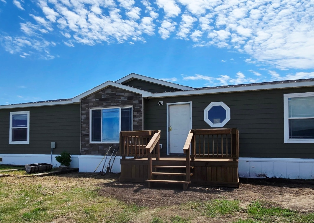 A single-story modular home with olive green siding, a stone accent wall, and a wooden front deck under a blue sky.