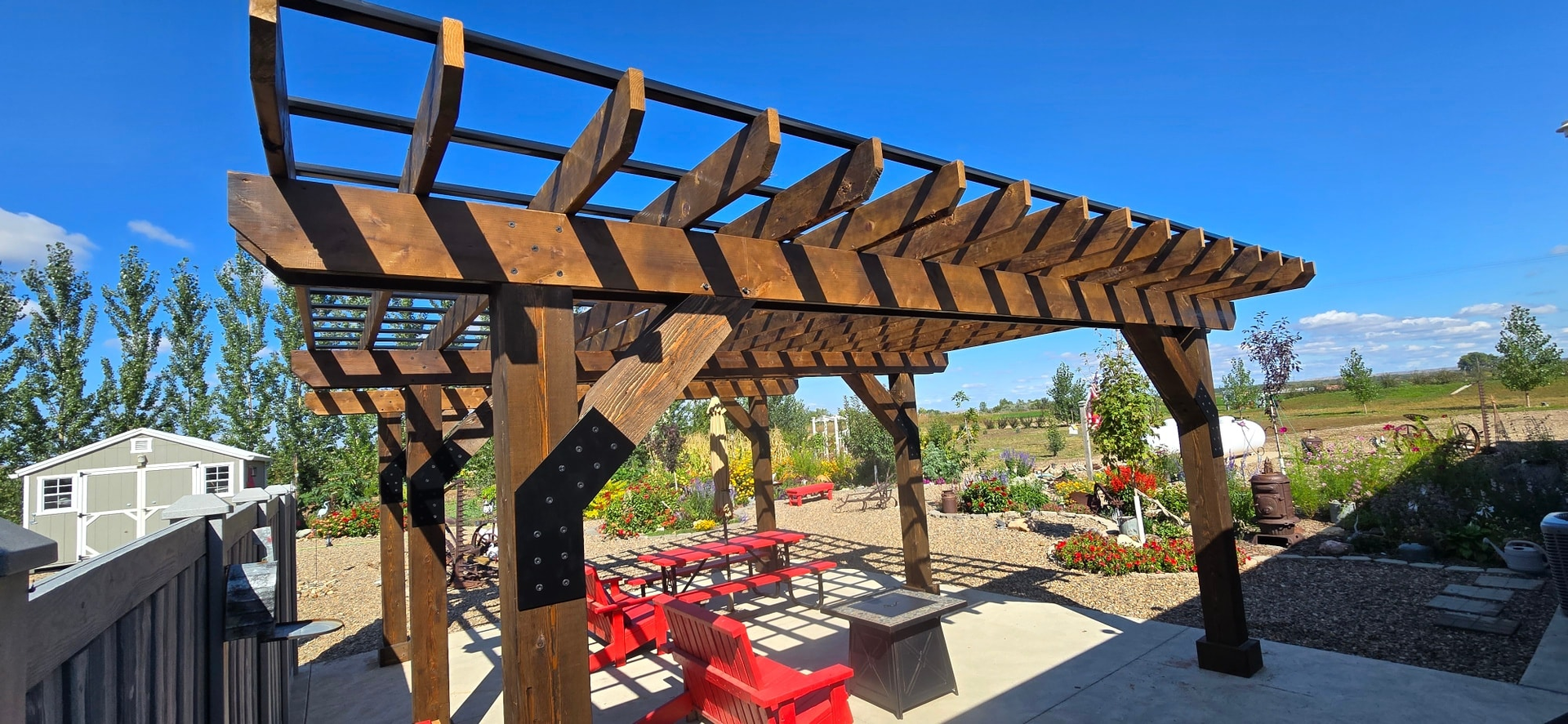 A wooden pergola with black metal brackets stands over a patio with red chairs on a sunny day.