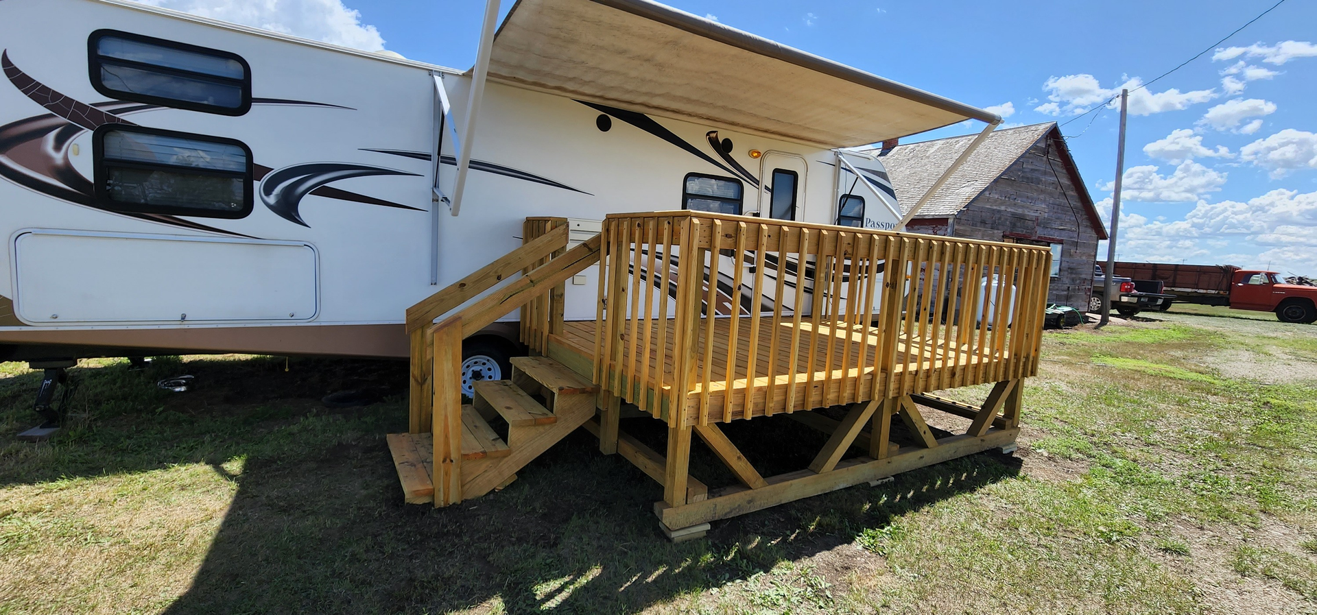 A new wooden deck and stairs built outside a camper trailer in an open, grassy field under a sunny blue sky.