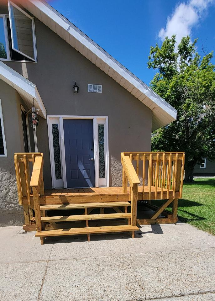 A new wooden deck and set of stairs leading to the front door of a gray house under a clear blue sky.