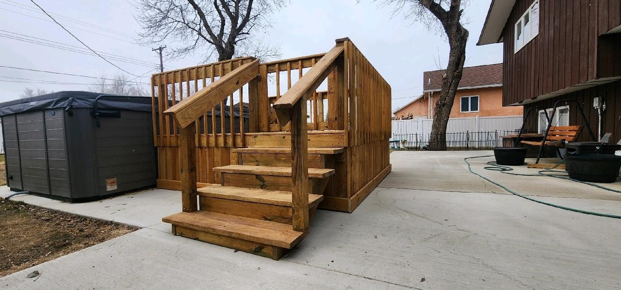 A wooden deck with a staircase and railing situated on a concrete patio next to a gray hot tub in a residential yard.