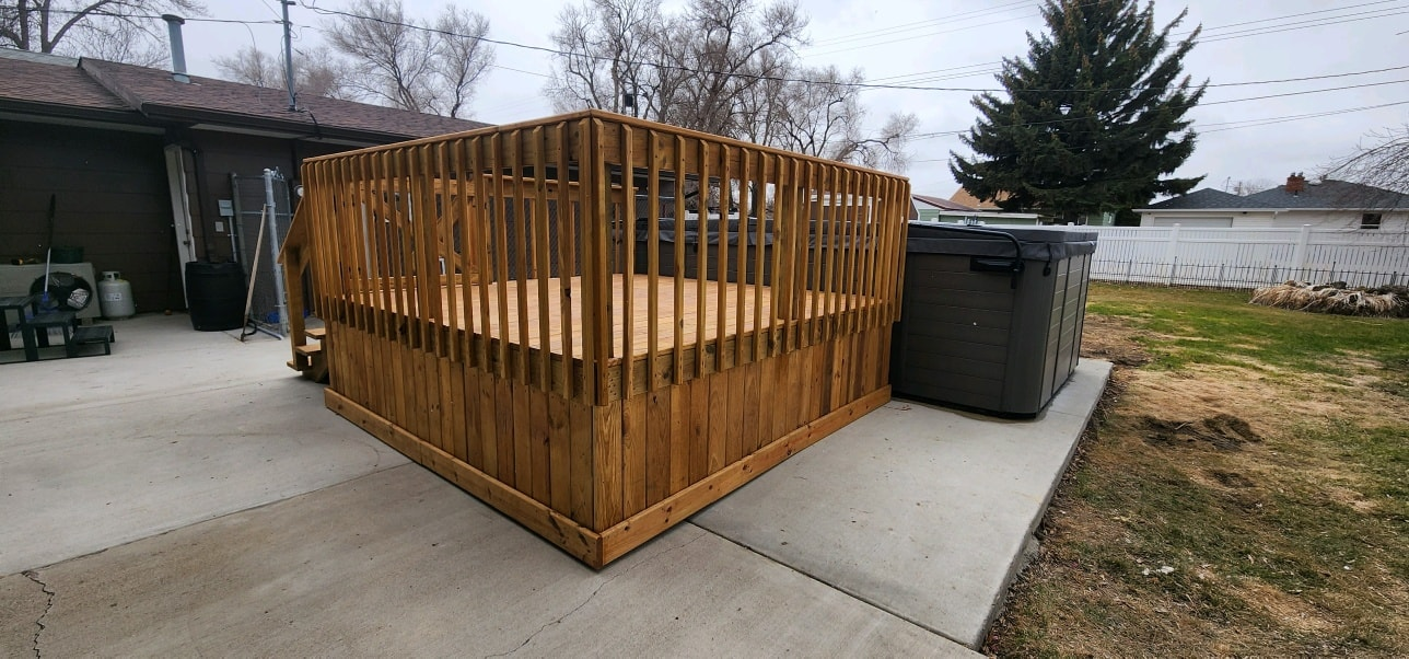 A wooden square deck with vertical railings sits on a concrete patio next to a large grey trash bin.