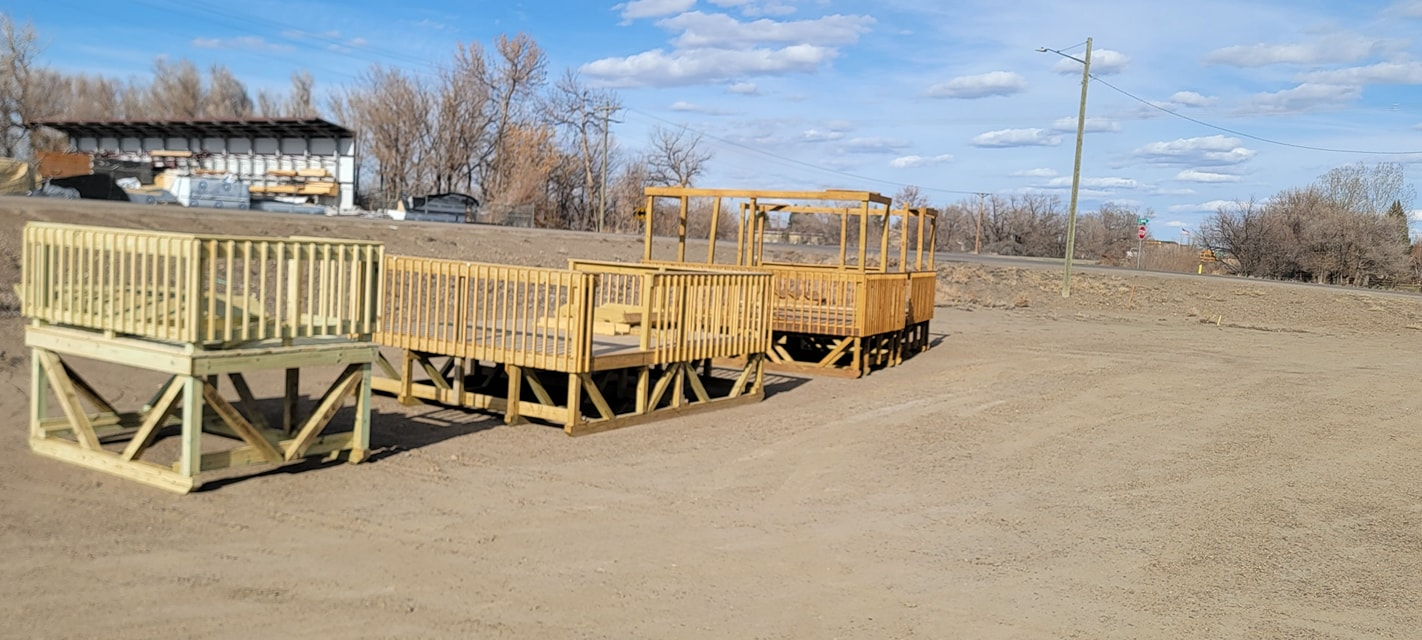 Several unfinished wooden hay feeders sit on a dirt lot with bare trees and a building in the background under a blue sky.