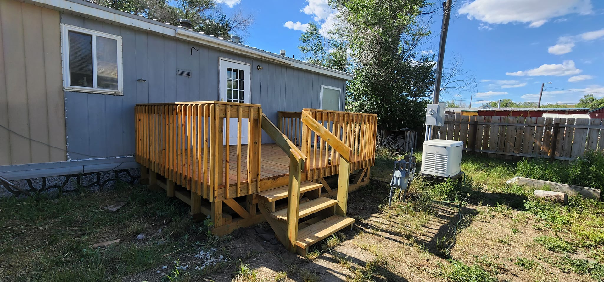 A light blue mobile home with a freshly built wooden deck and stairs, situated in a yard on a sunny day.